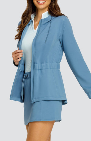 Model wearing a steel blue hooded jacket with a cinched waist and drawstring hood. Paired with a matching skort with drawstring waistband, and a light blue top with white accents.